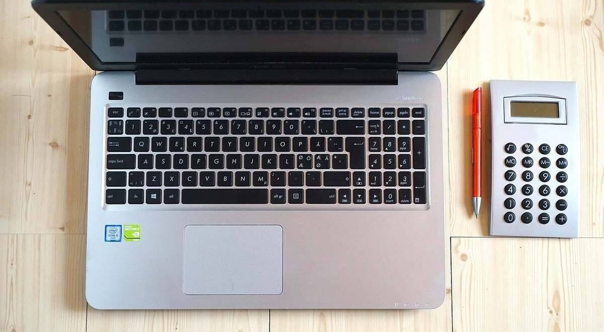 Ordinateur portable gris ouvert à côté d'une calculatrice de bureau blanche et d'un stylo orange sur bois