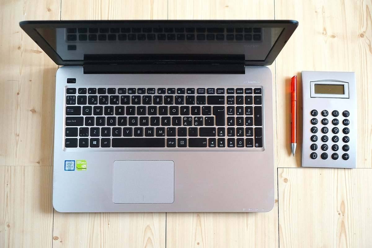 Ordinateur portable gris ouvert à côté d'une calculatrice de bureau blanche et d'un stylo orange sur bois
