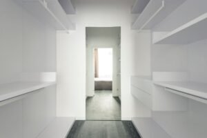 Corridor avec étagères blanches, sol gris, ouverture vers une pièce lumineuse avec lit