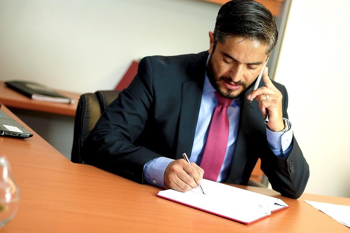 Homme d'affaires en costume travaillant à son bureau, écrivant sur un document tout en téléphonant Homme d'affaires en costume travaillant à son bureau, écrivant sur un document tout en téléphonant